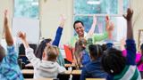 Multiracial group of children raising their hands in a classroom with a smiling Black woman teacher, photo by kali9/Getty Images