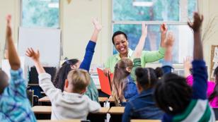 Multiracial group of children raising their hands in a classroom with a smiling Black woman teacher, photo by kali9/Getty Images