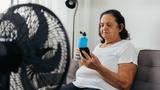 Elderly woman sitting on sofa in living room cooling off with floor fan, photo by klebercordeiro/Getty Images. 