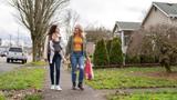 Two white women walking on the sidewalk in a neighborhood. One is carrying a baby in a carrier and the other is carrying grocery bags, Photo by Fly View Productions/Getty Images