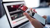 Closeup of a hand holding a cell phone showing a news website, with a laptop open in the background in soft focus, photo by oatawa/Getty Images