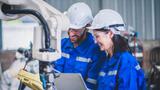 Two engineers in protective gear, one holding a laptop and the other looking at the controls for a robotic arm, photo by nuttawutnuy/Adobe Stock