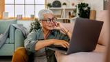 A senior woman wearing earbuds uses her laptop while sitting on the floor, photo by Pekic/Getty Images