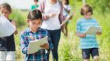 An elementary school girl writing in her notebook while outdoors with her class exploring nature, photo by FatCamera/Getty Images