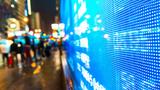 A lit display of stock market data showing on a busy city street at night, photo by baona/Getty Images