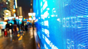 A lit display of stock market data showing on a busy city street at night, photo by baona/Getty Images