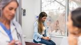A female doctor and the patient's mother talk unfocused in the foreground while a teen girl sits on the exam table looking upset, photo by SDI Productions/Getty Images