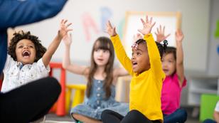 Diverse, smiling preschool students is sit on the floor in their classroom with their arms, raised  facing the teacher, photo by FatCamera/Getty Images