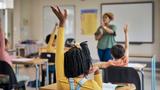 Teacher at the front of a classroom with students raising their hands, photo by pixdeluxe/Getty Images