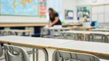 Chairs and desks in a classroom in focus with a teacher in the blurred background, photo by AJ_Watt/Getty Images