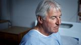 Elderly man in a hospital gown sitting on a hospital bed, photo by shapecharge/Getty Images