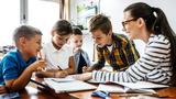 Elementary school students write in notebooks on a table with their teacher in front of them, photo by BalanceFormCreative/Adobe Stock