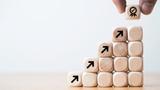 Two fingers placing the last block atop a stair-step stack of building blocks representing upward progress, photo by Dilok Klaisataporn/Getty Images