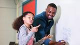 A Black male teacher works with a young female student in math class, photo by kali9/Getty Images
