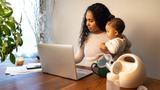 A young mixed-race African American mother holds her daughter while using a laptop, with a breast pump on the table, photo by Big Joe/Getty Images