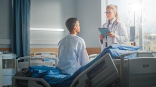 Young White man in a hospital gown sitting on a bed in a hospital talking to a white woman doctor, photo by gorodenkoff/Getty Images