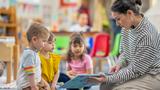 A preschool teacher sits on the floor of her classroom with a small group of students as she reads them a book.  The children are each dressed casually and are focused on the story.