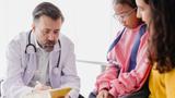 Doctor writing on a clipboard while talking with a mother and child, photo by coffeekai/Getty Images