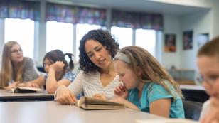A young ethnic teacher kneels by the desk of one of her students. The girl is reading an assignment in a book and sounding out words.