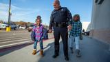 A police officer and two children stroll together in a parking lot, smiling, with various vehicles parked around them.