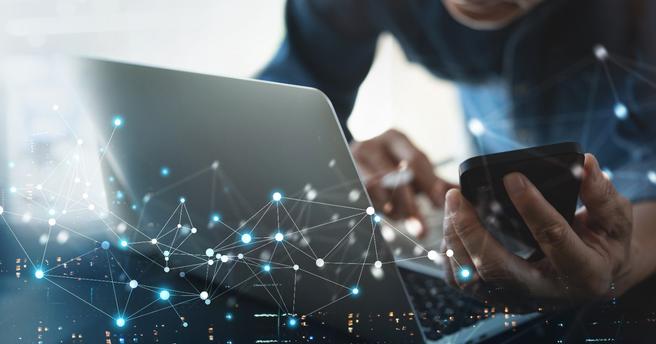 Person simultaneously working on a laptop and smartphone, with images representing the Internet of Things layered on top of the image, photo by tippapatt/Adobe Stock