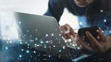 Person simultaneously working on a laptop and smartphone, with images representing the Internet of Things layered on top of the image, photo by tippapatt/Adobe Stock